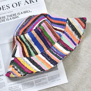 Op de afbeelding: Een veelkleurige gestreepte vissershoed met strepen in verschillende kleuren, waaronder roze, blauw, oranje en groen. De hoed is gemaakt van een textuurstof en ligt op een krant. De krantenkop luidt "THERE IS A WILL, THERE IS A WAY."