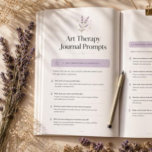 Pode incluir: Diário aberto com o título "Art Therapy Journal Prompts" e sugestões para autodescoberta e expressão emocional. Uma caneta repousa na página. Ramos de lavanda emolduram o diário.