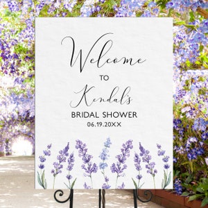 May include: White welcome sign with black calligraphy text that reads "Welcome to Kendall's Bridal Shower 06.19.20XX". The sign is decorated with a watercolor border of lavender flowers.