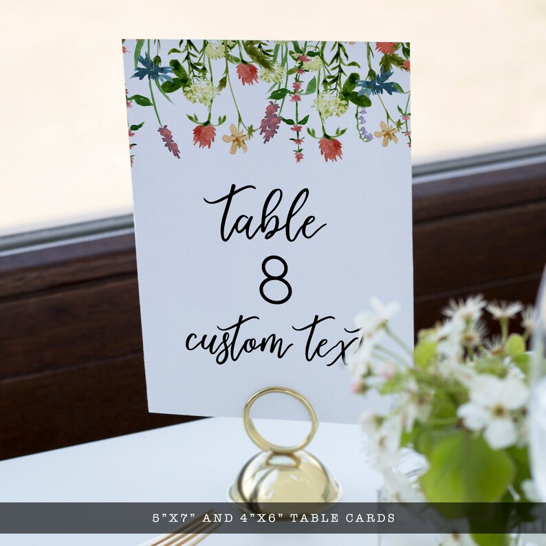 Pode incluir: Um cart&atilde;o de mesa branco com design floral em aquarela, exibindo "Table 8" e "custom text" em escrita preta. O cart&atilde;o &eacute; segurado por um suporte de metal dourado. A parte inferior do cart&atilde;o diz "5"X7" AND 4"X6" TABLE CARDS".