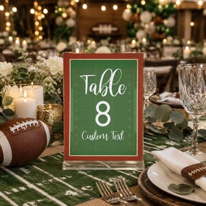 May include: A rectangular acrylic table sign featuring a green football field graphic with white yard lines. The text reads Table 8 and Custom Text in a white script and sans-serif font. The sign has a red border with a small white dot pattern.