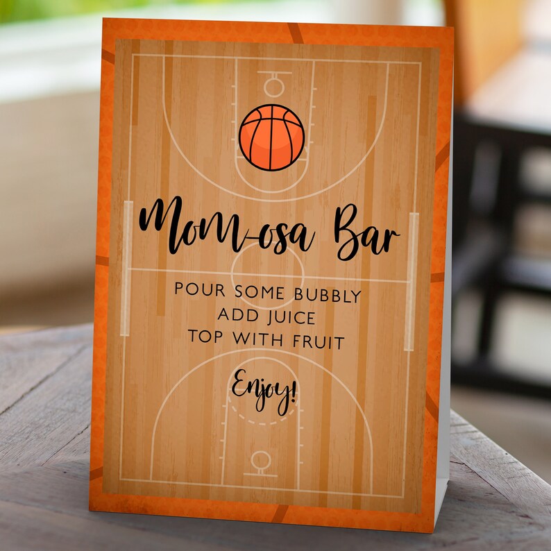 May include: A rectangular sign featuring a basketball court graphic with a wood grain pattern, an orange basketball icon, and black text that reads Mom-osa Bar, Pour some bubbly, Add juice, Top with fruit, and Enjoy! with an orange border.