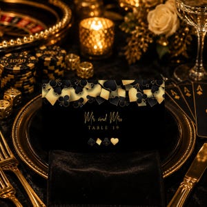 May include: A black rectangular table card featuring a gold and black playing card suit pattern at the top. The text reads Mr and Mrs TABLE 19 in gold script and serif font, with gold spade, diamond, club, and heart icons centred below.