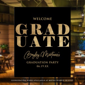 May include: A black graduation party sign with gold text. The sign reads "WELCOME GRADUATE Bayley Martinez's GRADUATION PARTY 06.19.XX". The sign is displayed on a metal stand.