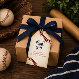 Può includere: Una scatola regalo quadrata in cartone marrone legata con un nastro blu scuro e un'etichetta color crema. L'etichetta presenta grafiche con cuciture da baseball rosse, il testo "Thank you" e "THANK YOU FOR SHOWERING ME WITH YOUR LOVE" in blu.