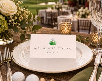 May include: A rectangular white place card with a golf ball and grass illustration, featuring the text MR. & MRS. YOUNG and TABLE 12, centered on a white plate with gold trim, surrounded by two golf balls, wooden tees, and silverware.