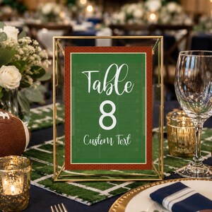 May include: A rectangular table number sign in a gold metal frame. The sign features a green football field design with white yard lines, the text "Table 8" in a white script and block font, and the words "Custom Text" in white script.