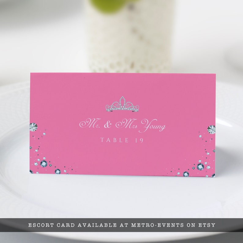 May include: Pink escort card with a silver tiara and the text "Mr. & Mrs. Young" and "Table 19" printed in white.