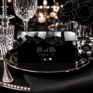 Op de afbeelding: Een rechthoekige zwart-witte tafelkaart met een casinothema en speelkaartsymbolen zoals schoppen, klaveren, ruiten en harten. De tekst luidt Mr and Mrs TABLE 19 in een wit script en schreeflettertype tegen een donkere achtergrond.