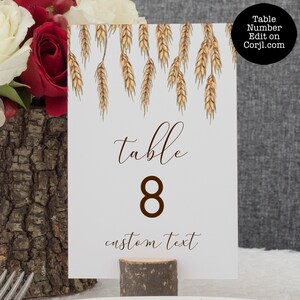 May include: White table number card with a brown wheat design at the top. The card says "table 8" in brown text and "custom text" in brown cursive text.