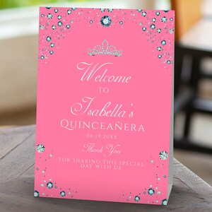 May include: Pink welcome sign with silver glitter and a silver crown. The sign says "Welcome to Isabella's Quincea&ntilde;era 06.19.20XX Thank You for sharing this special day with us".