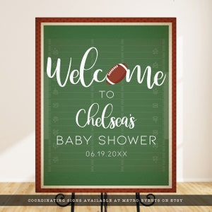 May include: A rectangular sign with a green football field background, white yard line markings, and a brown football graphic. The text reads: Welcome to Chelsea's BABY SHOWER 06.19.20XX. The sign has a dark red border with a small white dot pattern.