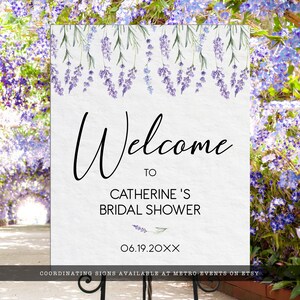 May include: A rectangular white sign featuring a watercolor illustration of purple lavender sprigs hanging from the top edge. The text reads Welcome to CATHERINE'S BRIDAL SHOWER and 06.19.20XX in black lettering. The sign is mounted on a black metal stand.