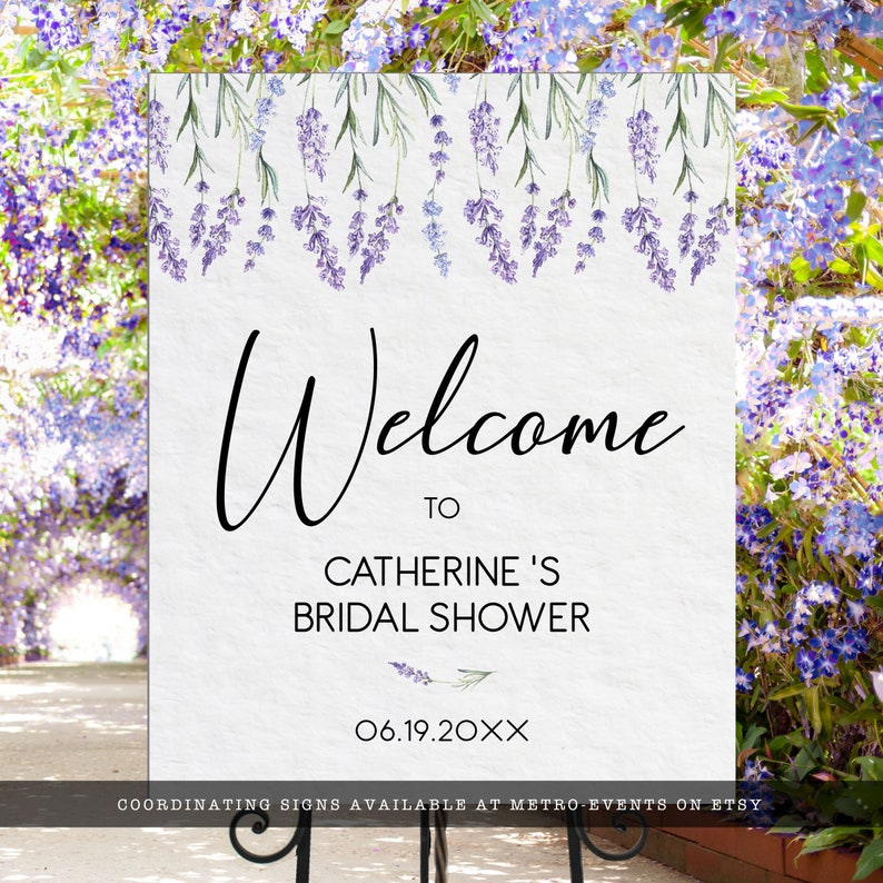 May include: A rectangular white sign featuring a watercolor illustration of purple lavender sprigs hanging from the top edge. The text reads Welcome to CATHERINE'S BRIDAL SHOWER and 06.19.20XX in black lettering. The sign is mounted on a black metal stand.