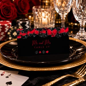 May include: A black rectangular table card featuring a red and black playing card suit pattern along the top edge. The text reads Mr and Mrs TABLE 19 in red script and block lettering, with small red and white suit icons at the bottom center.