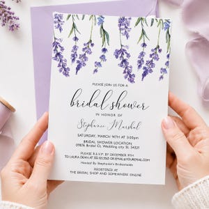 May include: A white bridal shower invitation with watercolor lavender sprigs. The invitation includes the text "Please join us for a bridal shower in honor of Stephanie Marshal" and event details. The invitation is held in a lavender envelope.