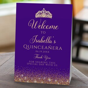 May include: A purple and gold Quincea&ntilde;era welcome sign. The sign features the text "Welcome to Isabella's Quincea&ntilde;era" with a gold tiara and glitter accents. The date is printed below the name, and a thank you message is at the bottom.