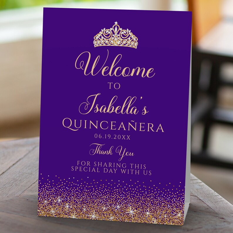 May include: A purple and gold Quincea&ntilde;era welcome sign. The sign features the text "Welcome to Isabella's Quincea&ntilde;era" with a gold tiara and glitter accents. The date is printed below the name, and a thank you message is at the bottom.