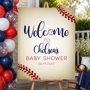 May include: A baseball-themed baby shower welcome sign on a wooden easel. The sign features the words "Welcome to Chelsea's Baby Shower" in navy and red, with baseball stitching accents. Balloons in red, white, and blue are on the left.