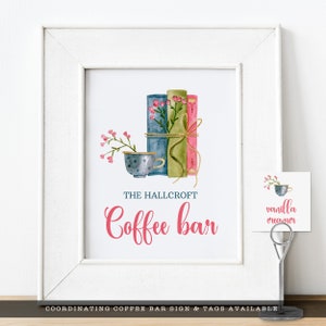 May include: A white framed print featuring a watercolor illustration of three stacked books in blue, green, and pink, tied with twine and topped with pink flowers next to a polka-dot teacup. The text reads THE HALLCROFT Coffee bar.
