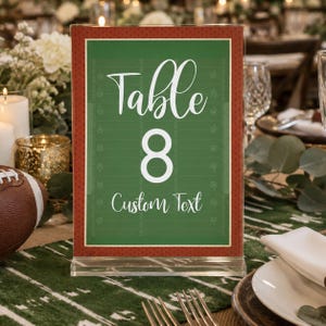 May include: A rectangular clear acrylic table sign featuring a green football field graphic with white yard line markings. The text reads Table 8 and Custom Text in white script and sans-serif fonts, framed by a red border with white dots.