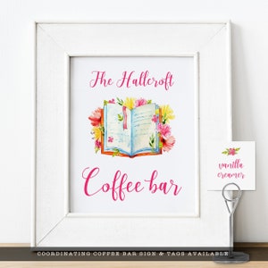 May include: A white framed sign with the text "The Hallcroft Coffee Bar" in pink script. An open book with floral accents is pictured above the text. A small white tag with the text "vanilla creamer" is on the right side of the sign.