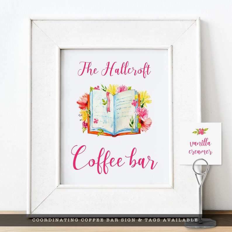 May include: A white framed sign with the text "The Hallcroft Coffee Bar" in pink script. An open book with floral accents is pictured above the text. A small white tag with the text "vanilla creamer" is on the right side of the sign.