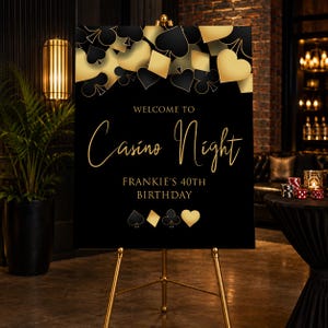 May include: A rectangular black sign on a gold easel featuring gold and black playing card suit symbols at the top and bottom. The text reads: WELCOME TO Casino Night FRANKIE'S 40TH BIRTHDAY. The design includes gold script lettering for the event title.