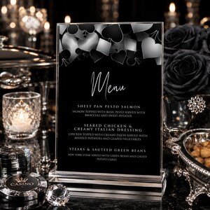 May include: A vertical rectangular menu card in a clear acrylic stand featuring a black background with silver playing card suit icons at the top. The text reads Menu, Sheet Pan Pesto Salmon, Seared Chicken & Creamy Italian Dressing, and Steaks & Sauteed Green Beans.