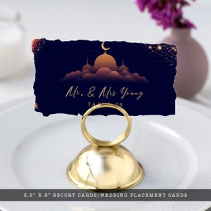 May include: A rectangular escort card with torn edges featuring a dark blue night sky, a gold crescent moon, and a mosque silhouette. The text reads Mr. & Mrs. Young Table 19. The card is held by a gold metal stand measuring 3.5 inches by 2 inches.