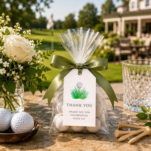 Può includere: Un sacchetto regalo in cellophane trasparente riempito con caramelle rotonde bianche, legato con un nastro di raso verde oliva. Un'etichetta rettangolare bianca mostra una pallina da golf su un tee nell'erba e il testo: THANK YOU THANK YOU FOR CELEBRATING WITH US!