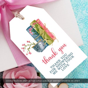 May include: A white rectangular favor tag with a watercolor illustration of three stacked books and a teacup with pink flowers. The text reads thank you and THANK YOU FOR SHOWERING ME WITH YOUR LOVE in pink and dark blue script and block lettering.