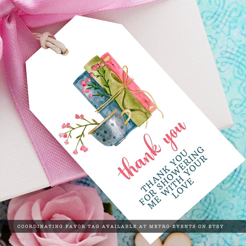 May include: A white rectangular favor tag with a watercolor illustration of three stacked books and a teacup with pink flowers. The text reads thank you and THANK YOU FOR SHOWERING ME WITH YOUR LOVE in pink and dark blue script and block lettering.