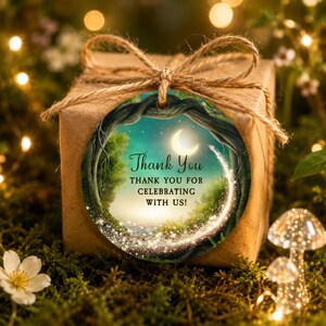 Op de afbeelding: Een rond cadeaulabel vastgebonden met bruin touw aan een vierkante doos. Het label bevat een cirkelvormige illustratie van een bos bij nacht met een maansikkel, gloeiende vonken en de tekst Thank You THANK YOU FOR CELEBRATING WITH US! in het zwart.