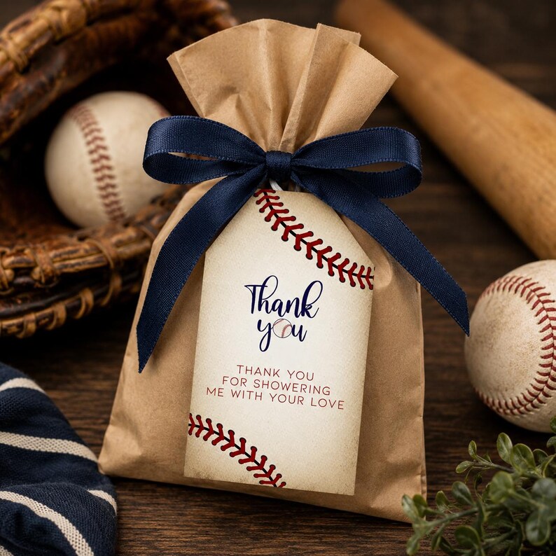 May include: A brown paper favor bag tied with a dark blue ribbon and a rectangular tag featuring a baseball stitch pattern. The tag displays the text Thank you and Thank you for showering me with your love in dark blue and red lettering.