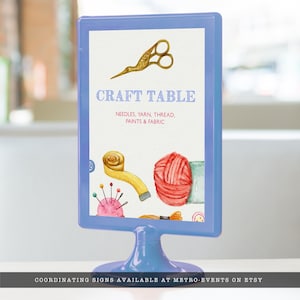 May include: A blue sign with the text "CRAFT TABLE" and a list of crafting supplies: needles, yarn, thread, paints, and fabric. The sign is decorated with watercolor illustrations of a pair of scissors, a spool of thread, a ball of yarn, and a pincushion.