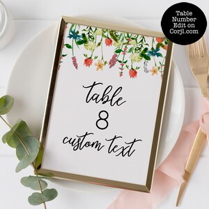 Pode incluir: Um cart&atilde;o de n&uacute;mero de mesa com moldura dourada com detalhes florais em aquarela. O cart&atilde;o exibe "Table 8" e "custom text" em uma escrita elegante. A parte superior do cart&atilde;o apresenta um design floral colorido com flores vermelhas, laranja e azul.