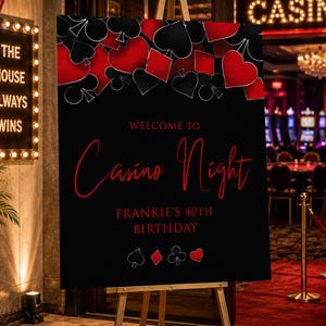 May include: A black rectangular sign on a wooden easel featuring red and white playing card suit symbols at the top and bottom. The text reads Welcome to Casino Night and Frankie's 40th Birthday in red and white lettering against a black background.