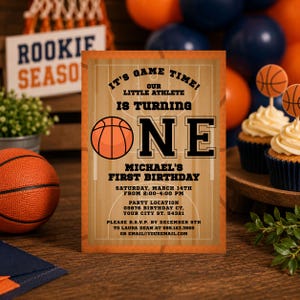 May include: A rectangular basketball-themed birthday invitation card featuring a wood court background, an orange basketball graphic, and black text that reads: IT'S GAME TIME! OUR LITTLE ATHLETE IS TURNING ONE, MICHAEL'S FIRST BIRTHDAY, SATURDAY, MARCH 14TH.