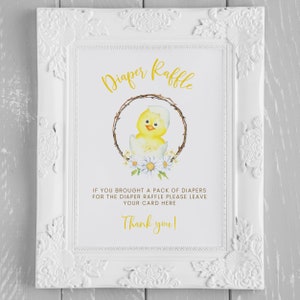 May include: White framed sign with the words "Diaper Raffle" in gold script. The sign features a watercolor illustration of a yellow chick hatching from an egg, surrounded by a wreath and daisies. Text below reads, "If you brought a pack of diapers for the diaper raffle please leave your card here. Thank you!"