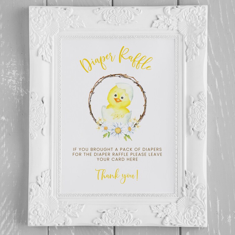 May include: White framed sign with the words "Diaper Raffle" in gold script. The sign features a watercolor illustration of a yellow chick hatching from an egg, surrounded by a wreath and daisies. Text below reads, "If you brought a pack of diapers for the diaper raffle please leave your card here. Thank you!"