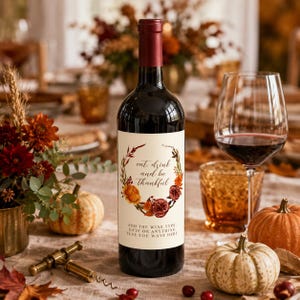 May include: A dark glass wine bottle with a cream-coloured label featuring a floral wreath illustration in autumn colours and the text "eat, drink and be thankful" followed by "ADD THE WINE TYPE, DATE OR ANYTHING ELSE, YOU WANT HERE".