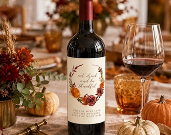 May include: A dark glass wine bottle with a cream-colored label featuring a floral wreath illustration in autumn colors and the text "eat, drink and be thankful" followed by "ADD THE WINE TYPE, DATE OR ANYTHING ELSE, YOU WANT HERE".