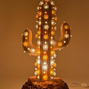 May include: A ceramic cactus lamp with a glossy brown finish, featuring two side arms and a textured base. The cactus is decorated with numerous small, translucent flower-shaped bulbs that emit a warm, glowing light throughout the structure.
