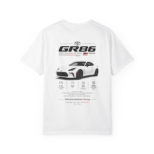 Peut inclure: T-shirt blanc avec un graphique d'une voiture de sport Toyota GR86 blanche. Le design comprend les spécifications de la voiture, le logo GR86 et le logo Toyota Gazoo Racing. Le texte se lit "DRIVEN BY BALANCE. BUILT FOR THE ROAD."