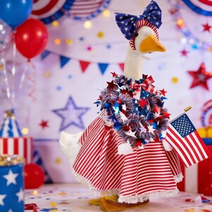 Puede incluir: Un ganso vestido con atuendo patriótico, que incluye un vestido a rayas rojas y blancas, un lazo con estrellas y una guirnalda de estrellas. Sostiene una pequeña bandera estadounidense, con un fondo festivo de globos y decoraciones.