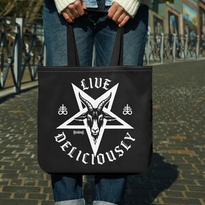 Puede incluir: Un bolso tote negro con un estampado gráfico blanco de una cabeza de cabra dentro de una estrella de cinco puntas. El texto "LIVE" aparece sobre la estrella, "DELICIOUSLY" debajo, y dos símbolos alquímicos de azufre a los lados.