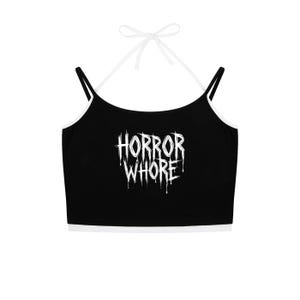 Puede incluir: Un top corto negro tipo halter con ribetes blancos y tirantes blancos finos que se atan al cuello. La parte delantera presenta el texto "HORROR WHORE" en una fuente blanca, goteante y dentada que imita la estética de las películas de terror.