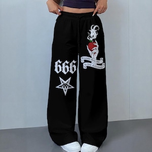 Puede incluir: Pantalones de chándal negros de pierna ancha con un gráfico blanco del número 666 y un pentagrama en la pierna izquierda. La pierna derecha muestra una ilustración blanca de una mano sosteniendo una manzana roja con una cabeza de cabra y el texto Wouldst thou like to live deliciously?