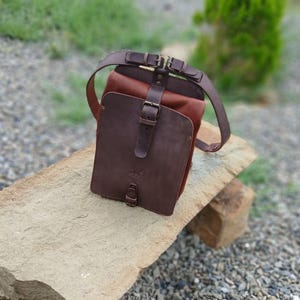 May include: A structured leather backpack featuring a dark brown front flap with a vertical strap and buckle closure. The main body is a lighter reddish-brown leather, complemented by dark brown adjustable shoulder straps and metal hardware.